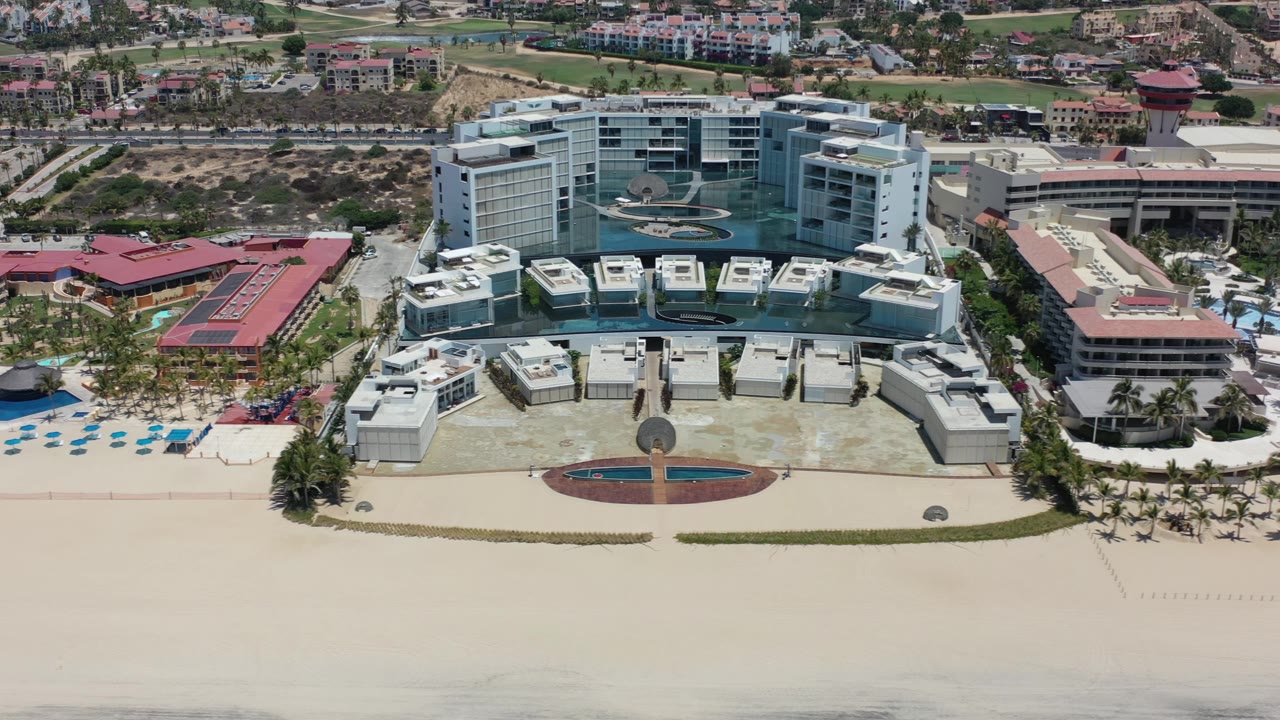 Elevated Los Cabos resort view with the main white hotel centered, a wide empty sand foreground, pool decks at left, and low buildings extending behind the property.
