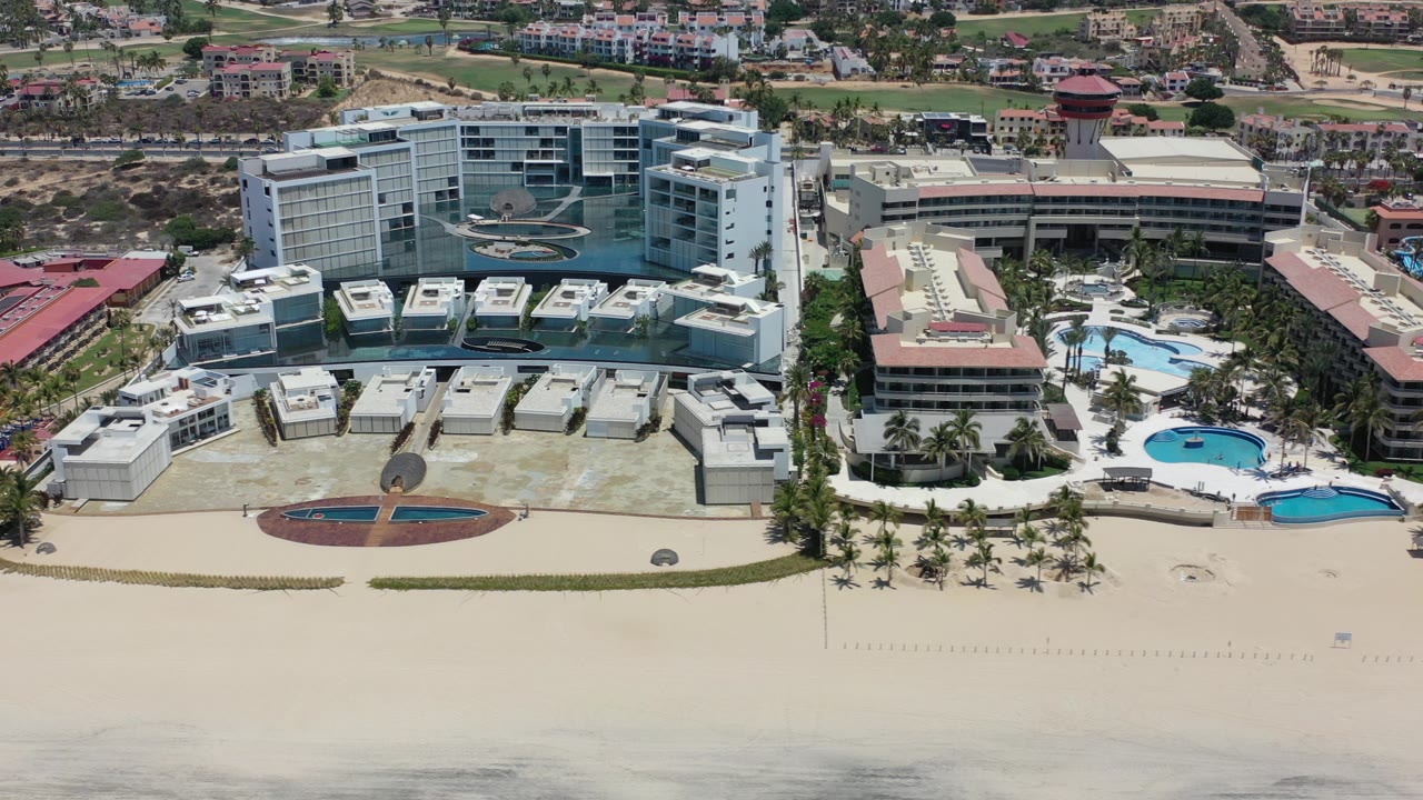 Elevated view in Los Cabos, Mexico showing beachfront high-rise resort buildings, palm-lined courtyards, swimming pools, pale sand, and no visible people at the opening frame.