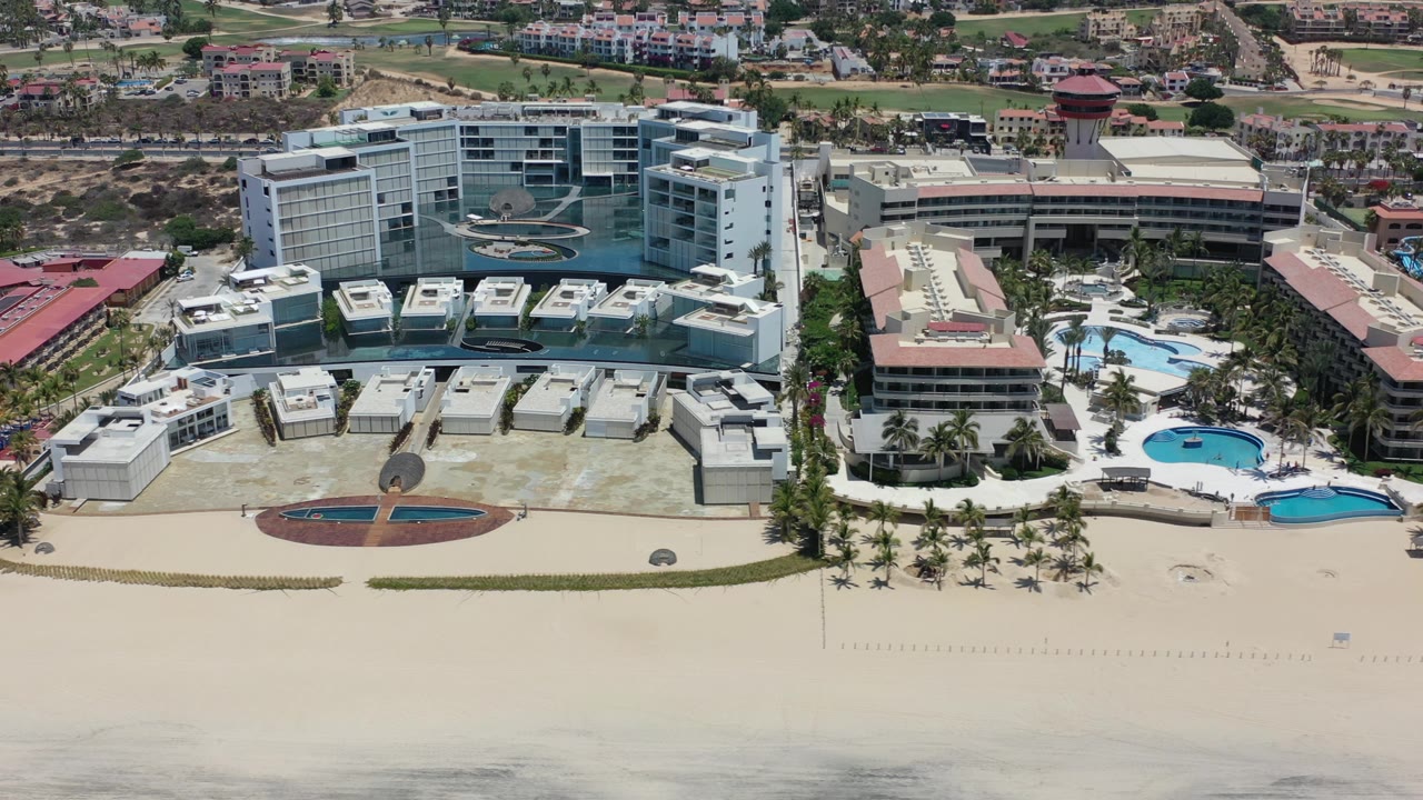 Aerial frame over Los Cabos, Mexico with the resort buildings centered, two blue pools to the right, rooftop details, open beach foreground, and no visible people.