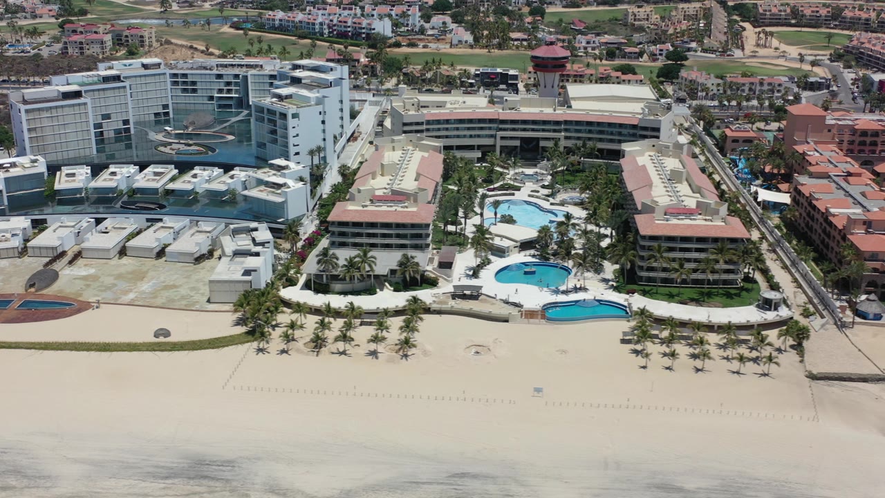 Aerial frame over Los Cabos with a broad sand foreground, resort pools near the center, a large white hotel on the left, terracotta buildings to the right, and no visible people.