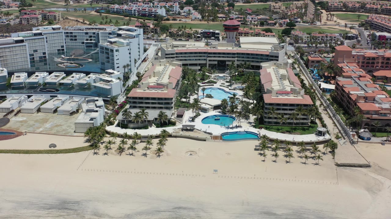 Aerial resort shoreline with hotels, pools, palms, and sand.