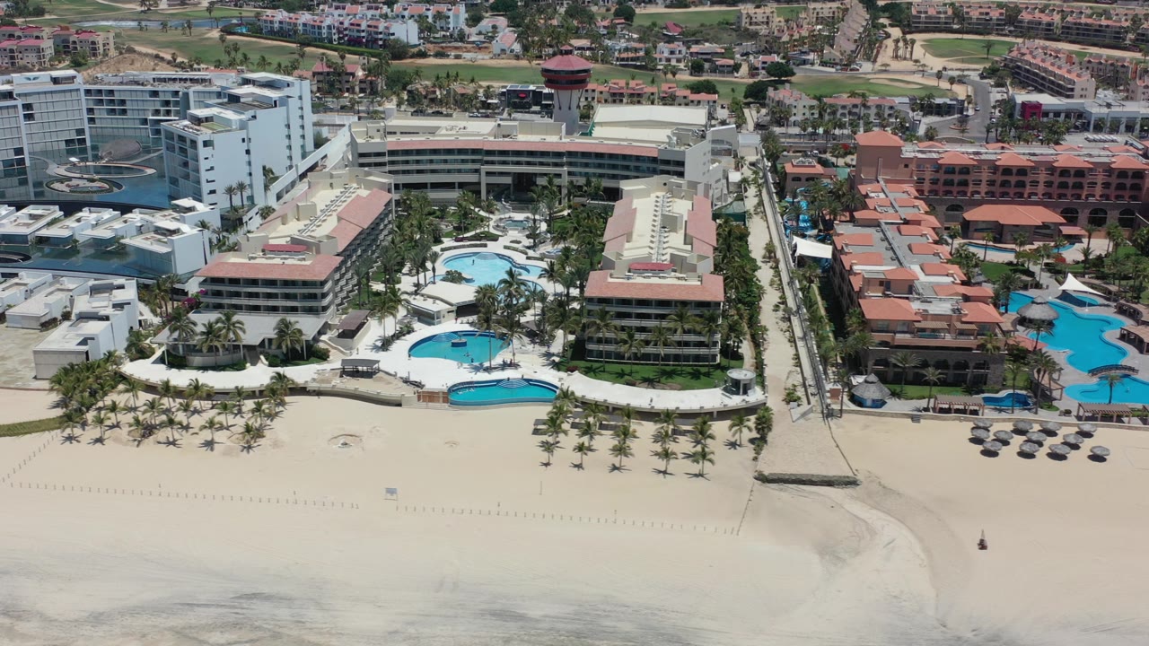 Aerial view of beachfront hotels, pools, palms, and pale sand.