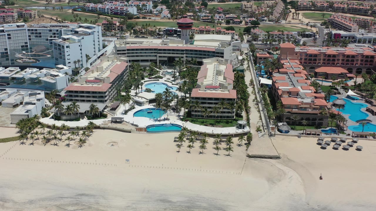 Aerial frame of the Los Cabos, Mexico hotel frontage showing curved blue pools, rows of palms, beach umbrellas, and broad sand in the foreground.