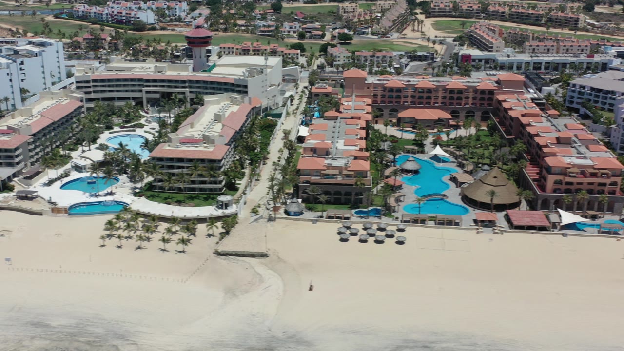 High aerial view over Los Cabos beachside resort buildings, blue pools, pale sand, and a thin surf line, with no visible people.