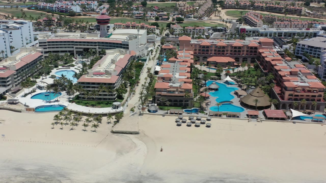Aerial frame over Los Cabos resorts with curved blue swimming pools, palm rows, broad sand in front, and no visible people.
