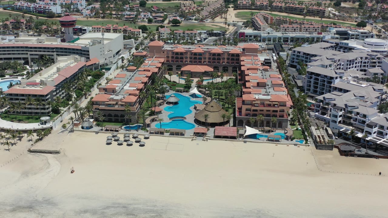 Aerial frame from Los Cabos, Mexico showing a beachfront hotel complex, turquoise pool areas, pale sand, low surf, and no visible people.