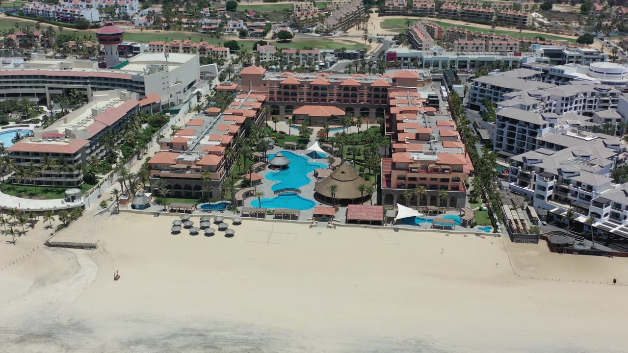 Aerial view in Los Cabos, Mexico with the beach foreground, resort pools centered, red-roof buildings to the right, white hotel structures to the left, and no visible people.