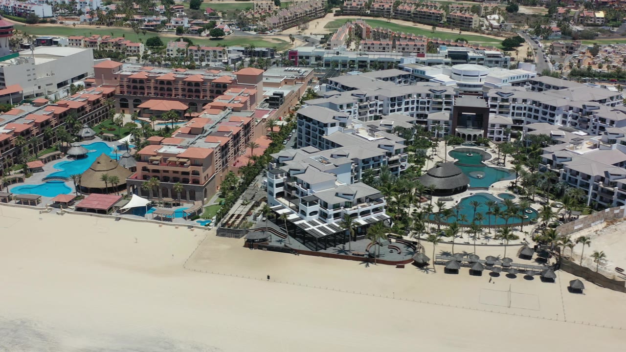 Aerial view in Los Cabos showing the resort pool complex more centered, with white and terracotta hotel buildings behind the sand beach and no visible people.