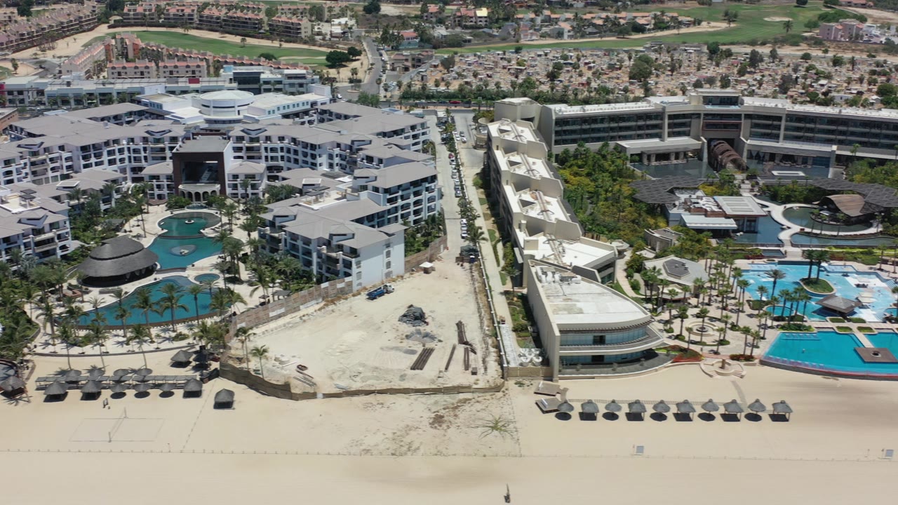 Aerial view of beachfront resort buildings, pools, umbrellas, sand, and inland roads.