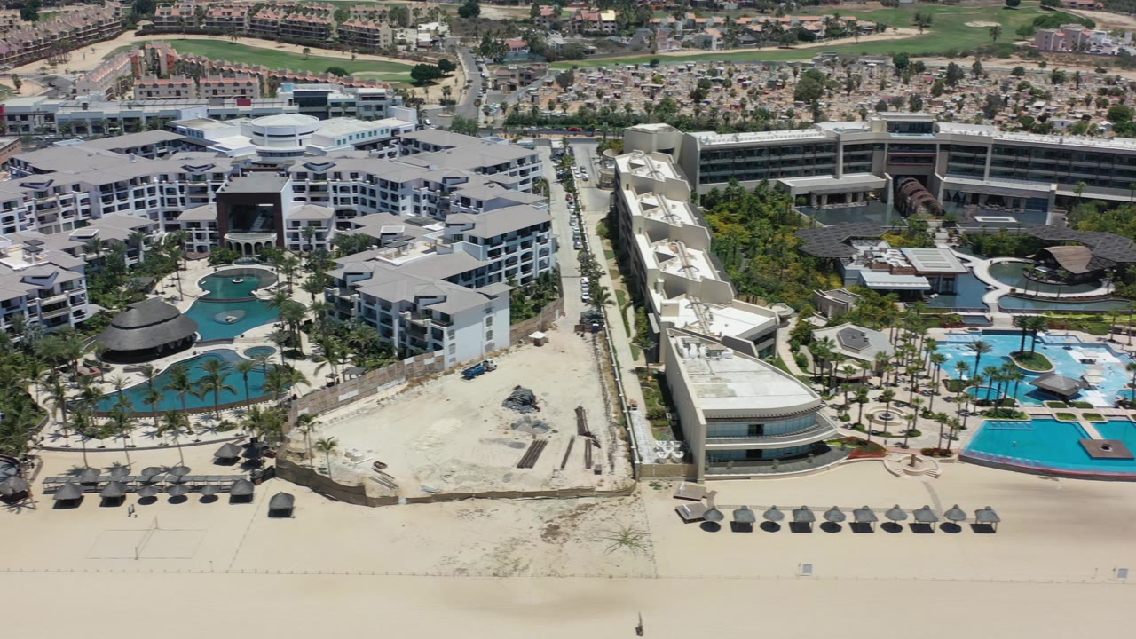 Los Cabos aerial view with a wider look at turquoise pool areas, white hotel blocks, beachfront sand, inland streets, and no visible people.