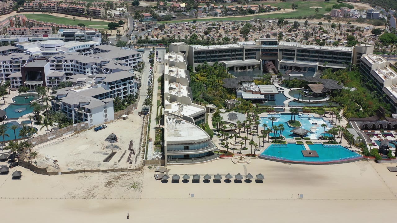 Los Cabos aerial view looking over white hotel roofs, a central pool area, palms, pale sand, inland roads, and no visible people.