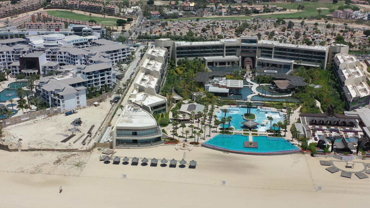Aerial view over a Los Cabos, Mexico beachfront resort, showing a wide sandy beach in front of turquoise pools, palm landscaping, and large hotel buildings with no identifiable people visible.