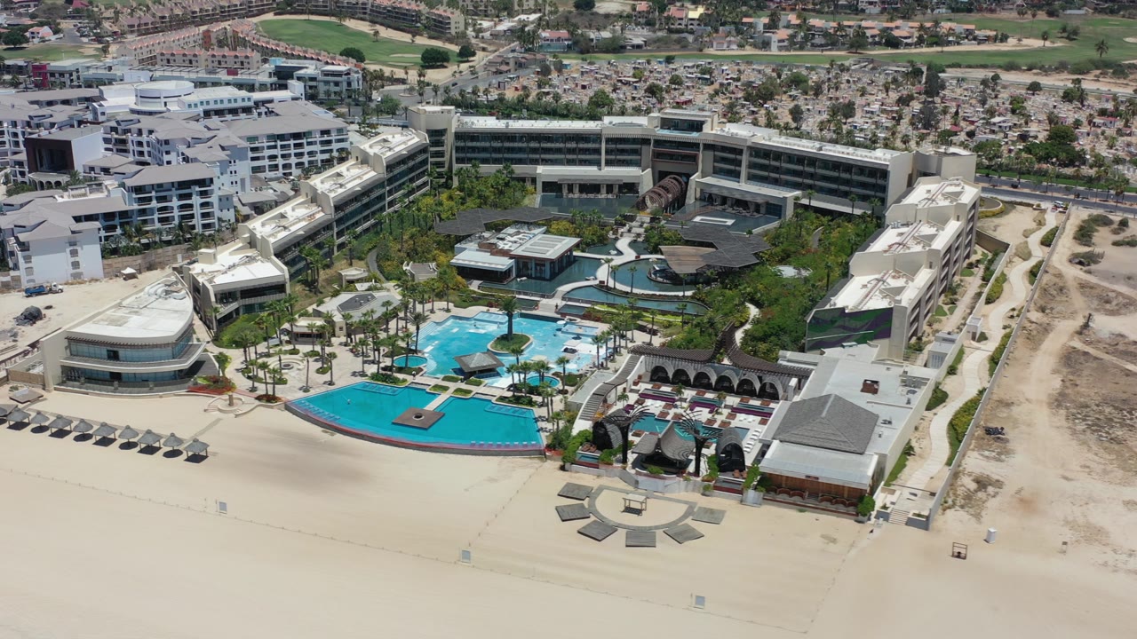 Aerial view over Los Cabos, Mexico showing a beachfront hotel complex, bright turquoise pools, pale sand, palms, and inland roads with no visible people.