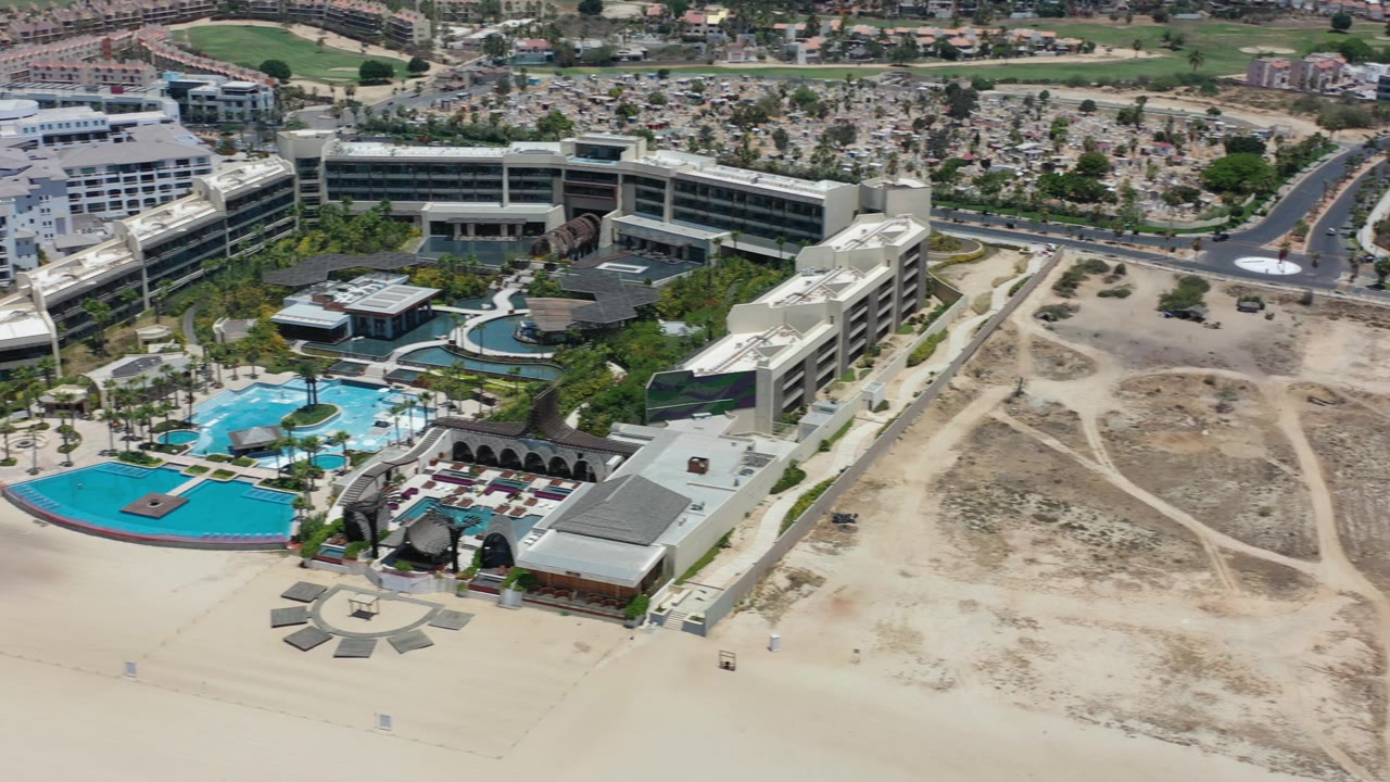 Oblique aerial view in Los Cabos, Mexico, showing a beach resort with bright blue pools beside a large empty sandy lot, roads, low-rise buildings, and no visible people.