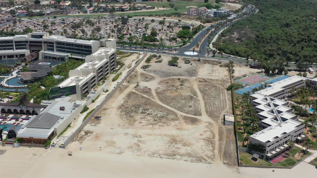 Opening aerial frame over a large sandy beachfront lot in Los Cabos, Mexico, with low-rise buildings, roads, vegetation, and no visible people.