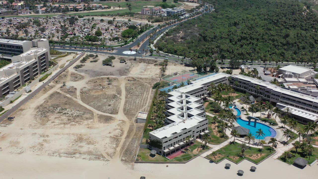 Aerial view of a beachfront building complex with a blue pool and sandy open land.