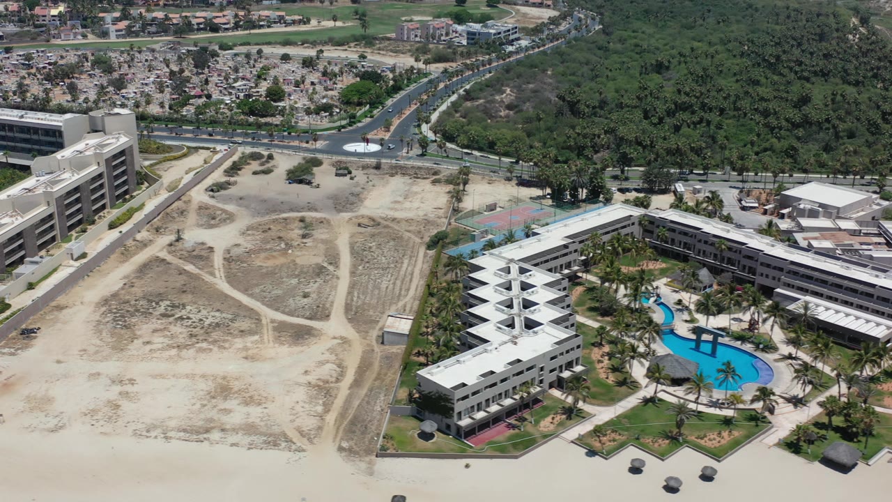 Aerial view in Los Cabos, Mexico, with the building complex centered lower in frame, a bright blue pool inside the landscaped grounds, sandy land to the left, and no visible people.