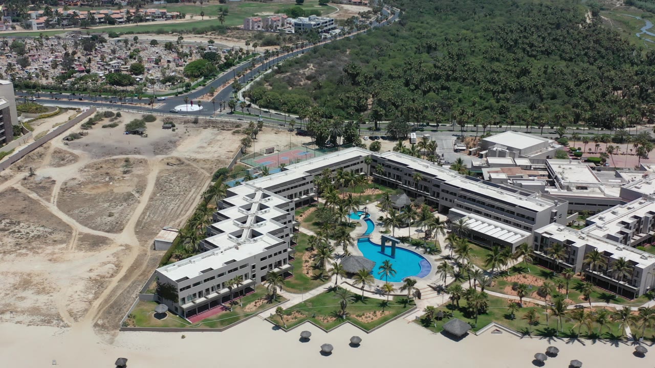 Opening aerial frame over Los Cabos shows a light-colored building complex with curved blue pools, palm landscaping, sandy open land, dense green forest, and no visible people.