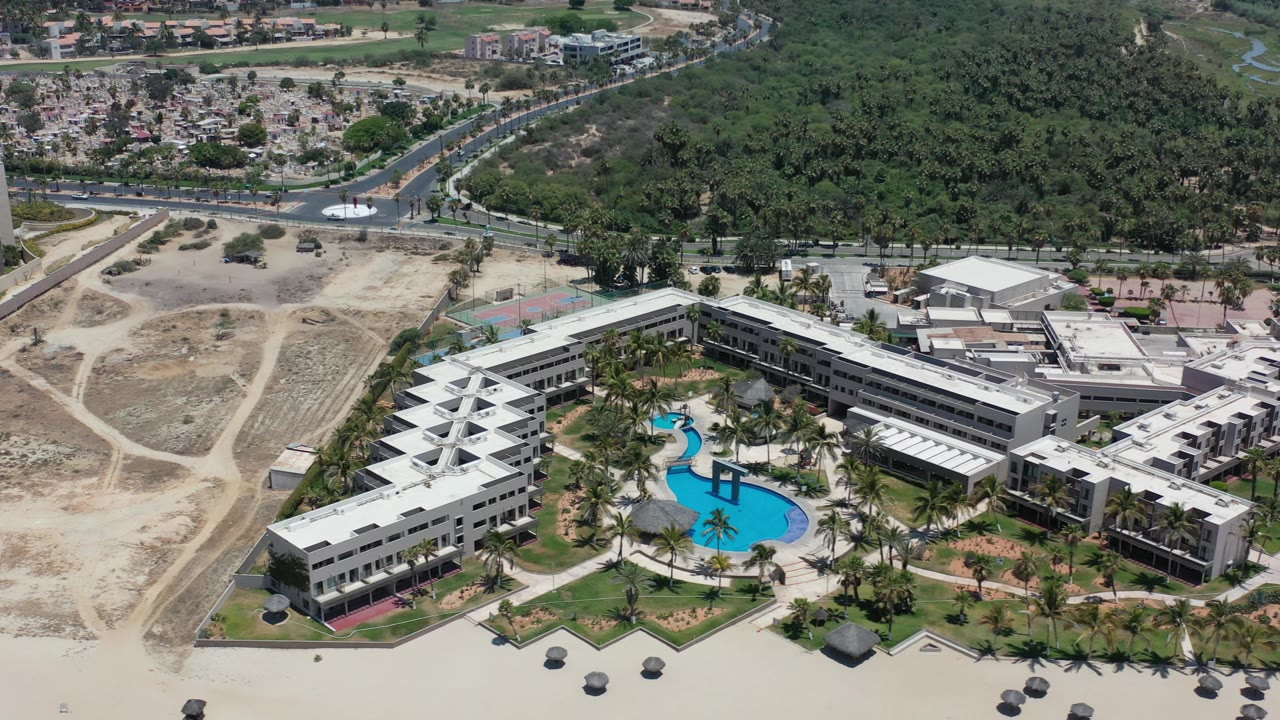Aerial shot of resort-like buildings, pool courtyard, sandy lots, road, and forest.