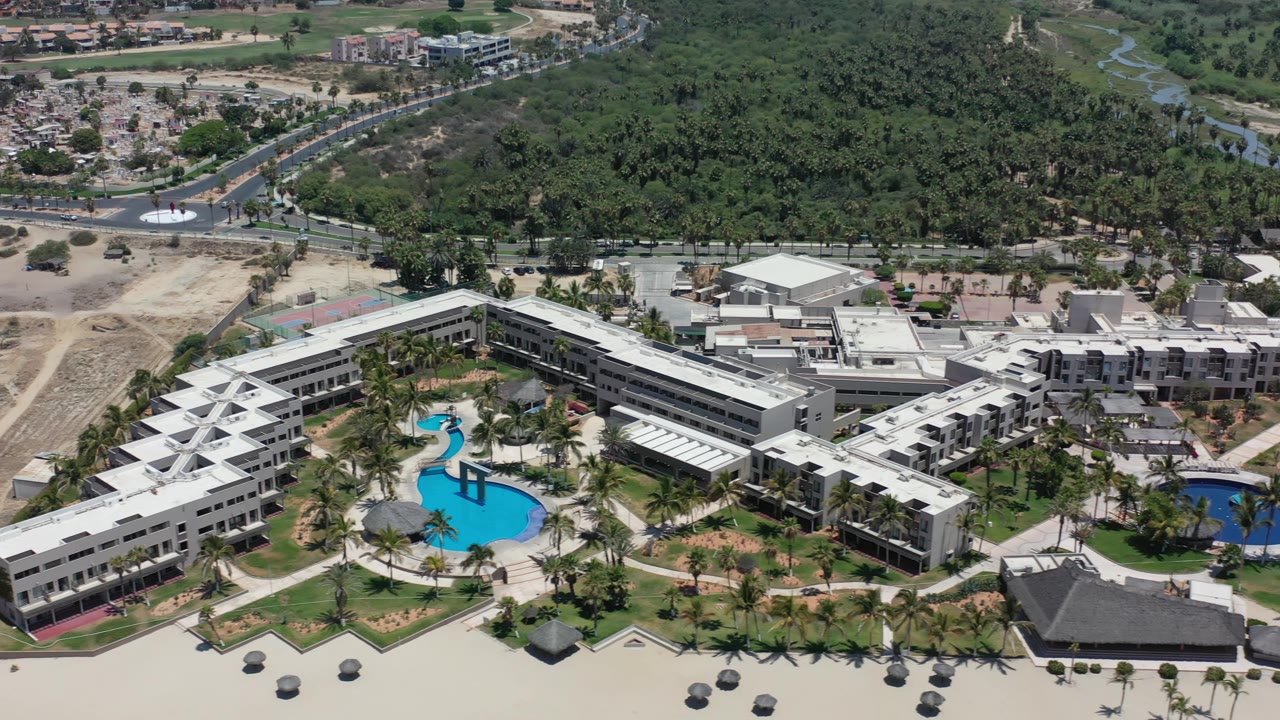 Aerial view in Los Cabos with the resort centered, a turquoise pool near the left wing, a darker pool to the right, palm rows along the sand, and no visible people.