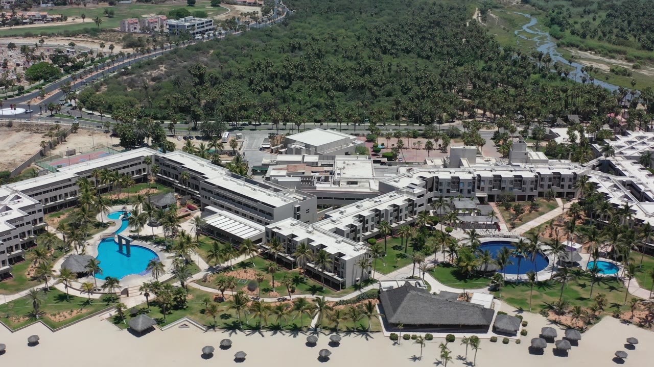 Opening aerial frame over Los Cabos, Mexico, showing a beachfront resort complex with bright blue pools, palm landscaping, sand umbrellas, roads, dense greenery, and no visible people.
