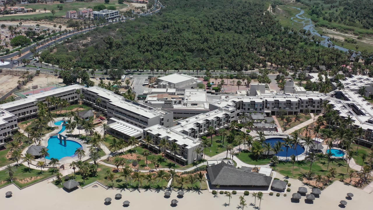High aerial of coastal hotel complex with pools, palms, beach, and green inland corridor.