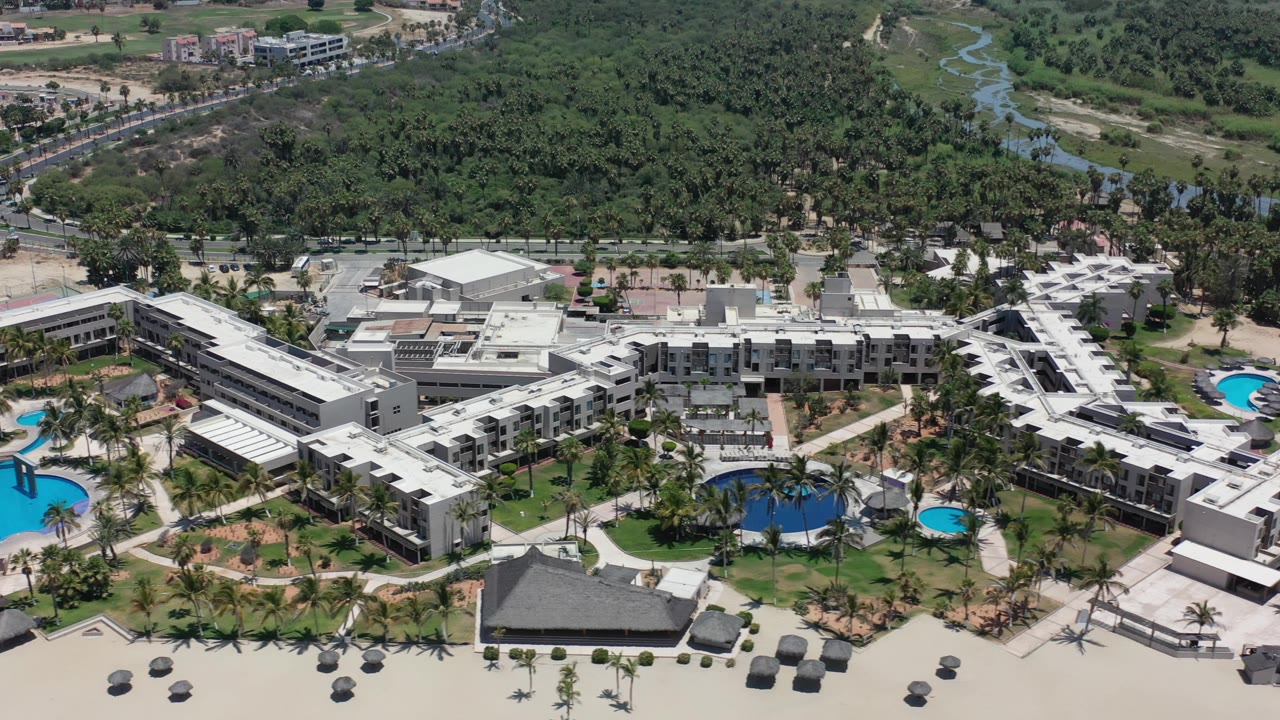 Aerial frame in Los Cabos, Mexico, with the building complex centered, multiple bright blue pools visible, palms along paths, dense greenery behind, and no visible people.