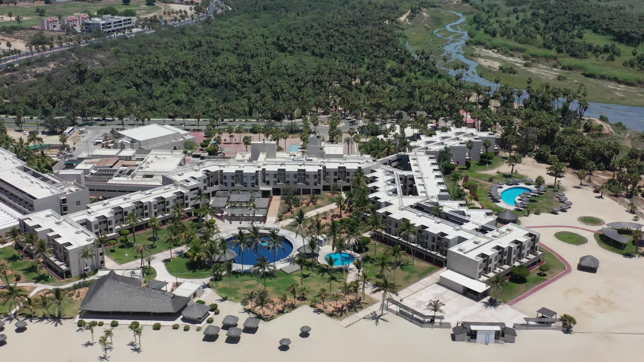 Aerial view of white resort buildings, pools, palms, sand, and green river terrain.