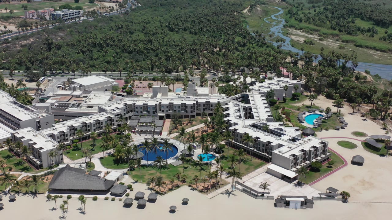 Aerial view in Los Cabos, Mexico with the white poolside buildings centered, blue swimming pools at the lower edge, sandy open areas, palms, and a green river corridor in the background, with no visible people.