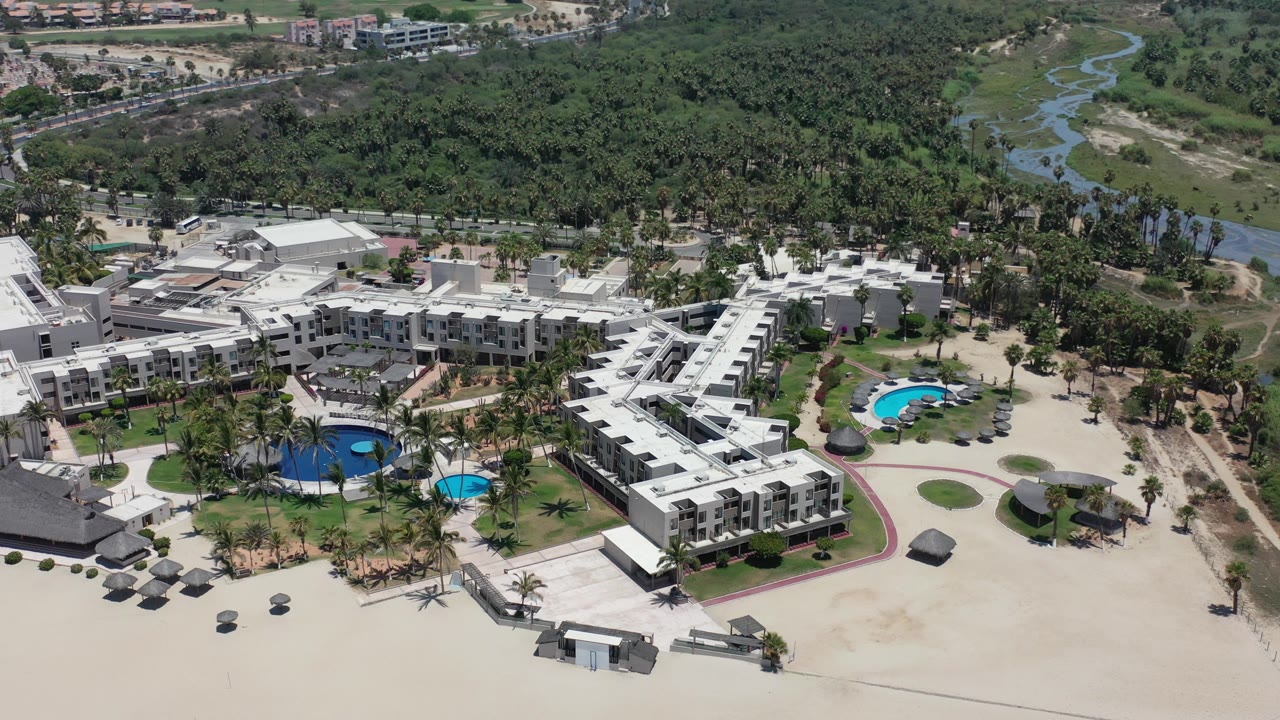 Aerial view of white beachfront buildings, pools, palms, sand, and green wetlands