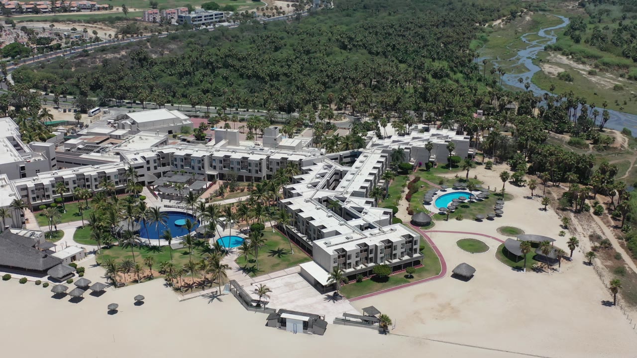 Aerial frame in Los Cabos, Mexico with the resort buildings centered, two bright blue pool areas below, beach umbrellas on the sand, and dense green vegetation behind the property.