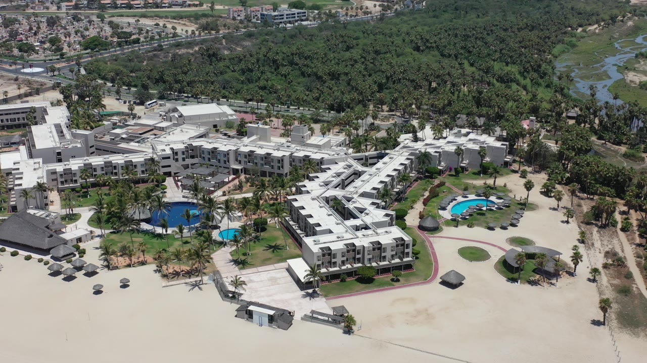 Aerial opening frame over Los Cabos, Mexico, showing white beachfront hotel buildings, pools, sandy foreground, dense green trees beyond, and no visible people.