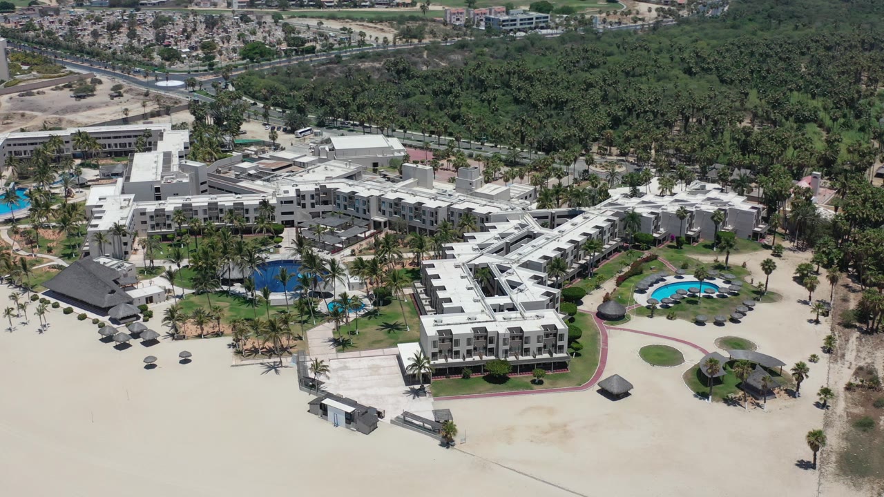 Aerial view of a large white beach resort with pools, palms, sand, and trees.