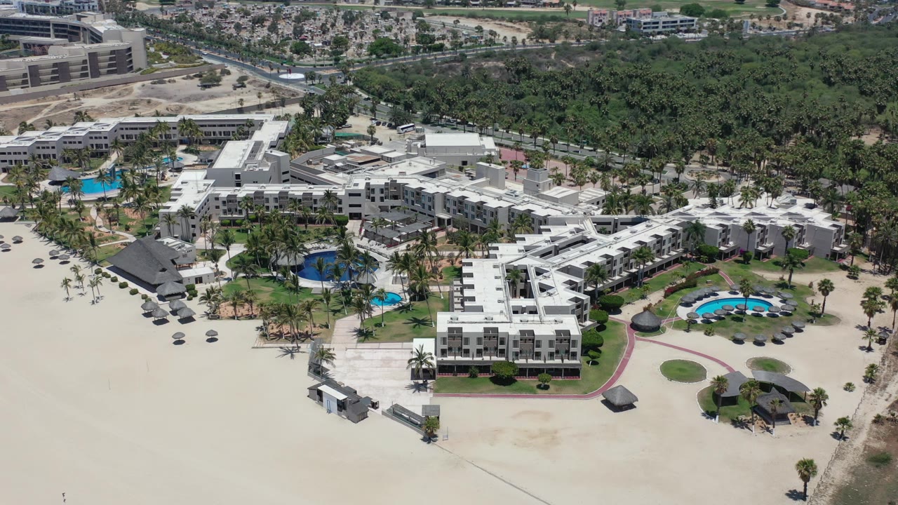 Aerial frame in Los Cabos, Mexico with the hotel centered, a circular pool area on the right, sand in the foreground, tree canopy behind, and no visible people.