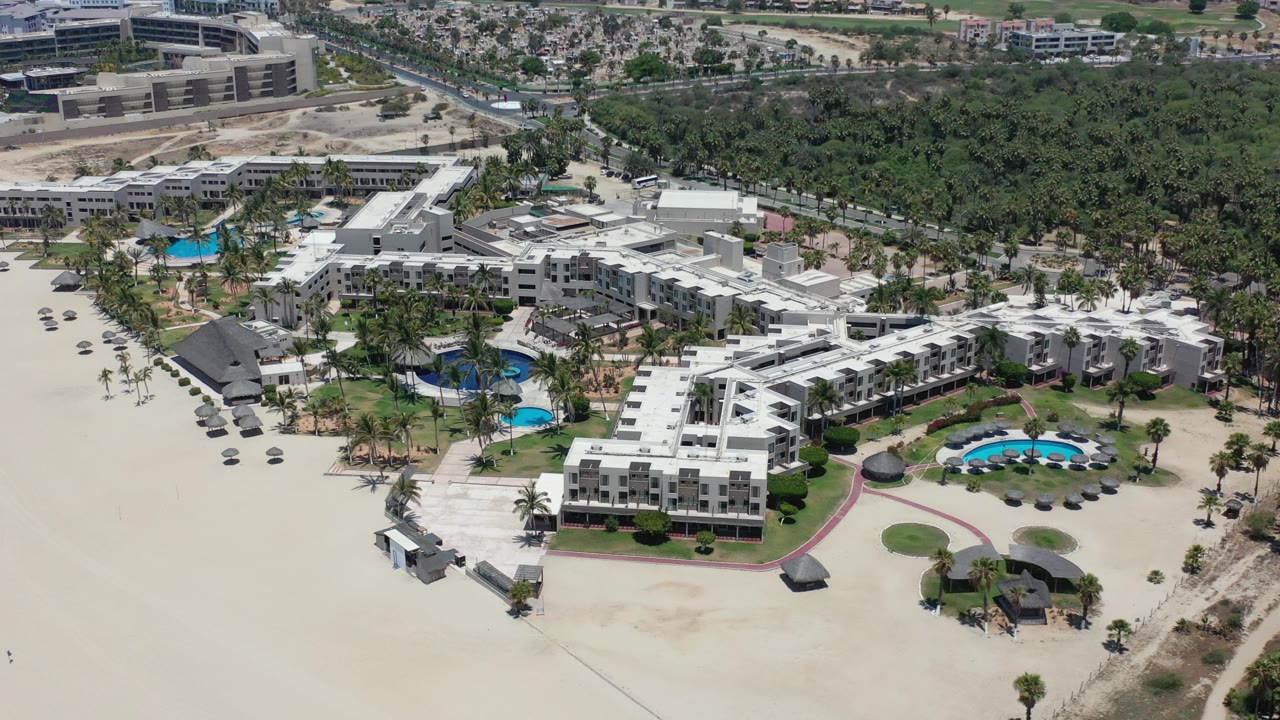 Aerial view of white beachfront buildings, pools, sand, palms, and dense trees.