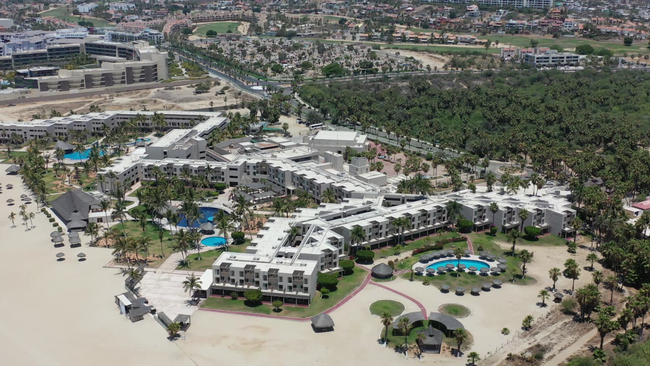 Opening aerial frame over Los Cabos, Mexico, showing white beachfront resort buildings, blue pools, pale sand, palm cover, and distant city blocks with no visible people.