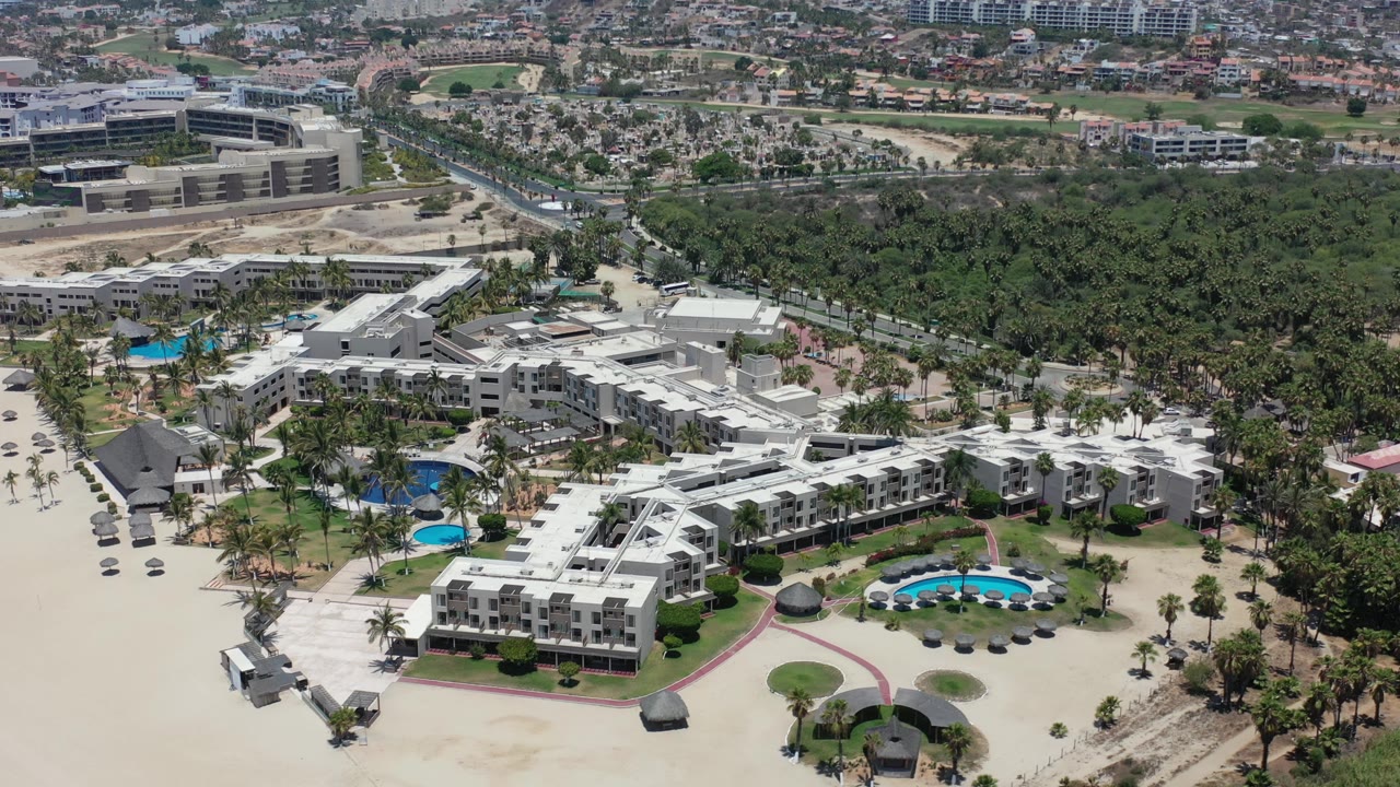 Drone view of resort pools, sandy beachfront grounds, palms, and surrounding development.