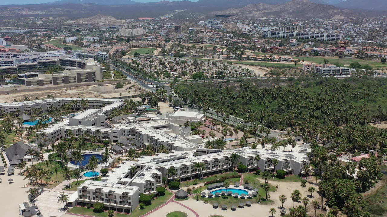 Opening aerial frame over Los Cabos with white resort buildings and blue pools in the foreground, a dense green palm area through the center, city blocks beyond, dry mountains on the horizon, and no visible people.