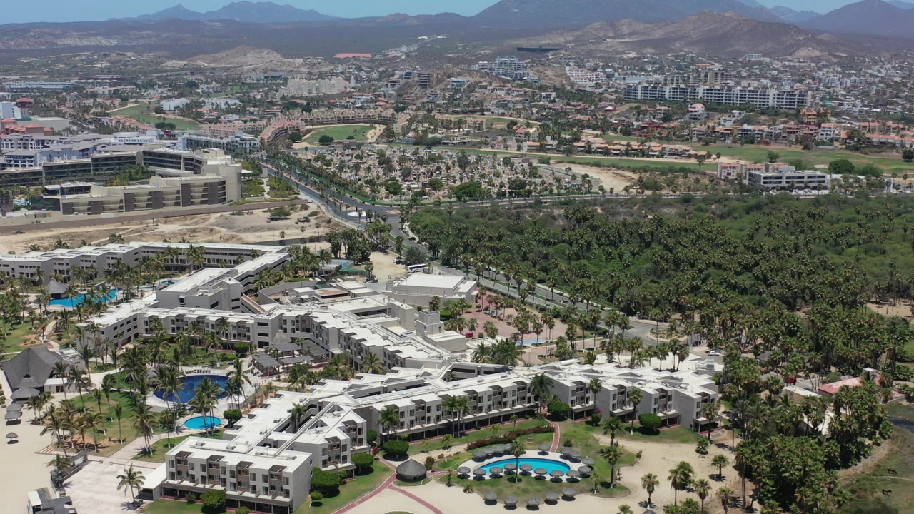Wide drone view of hotels, palm canopy, urban development, and mountain skyline.