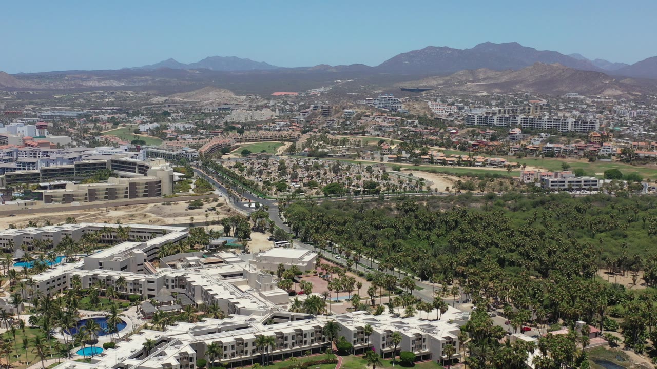 Wide aerial frame over Los Cabos, Mexico with white resort roofs along the bottom, a large green canopy through the center, roads and low-rise buildings beyond, brown mountains on the horizon, and no visible people.
