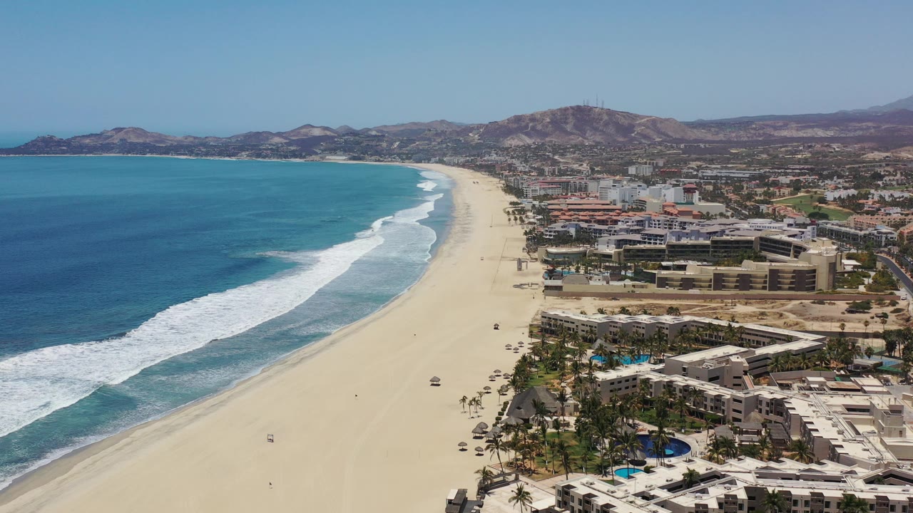 Aerial view of beach, ocean surf, resort buildings, and inland hills.
