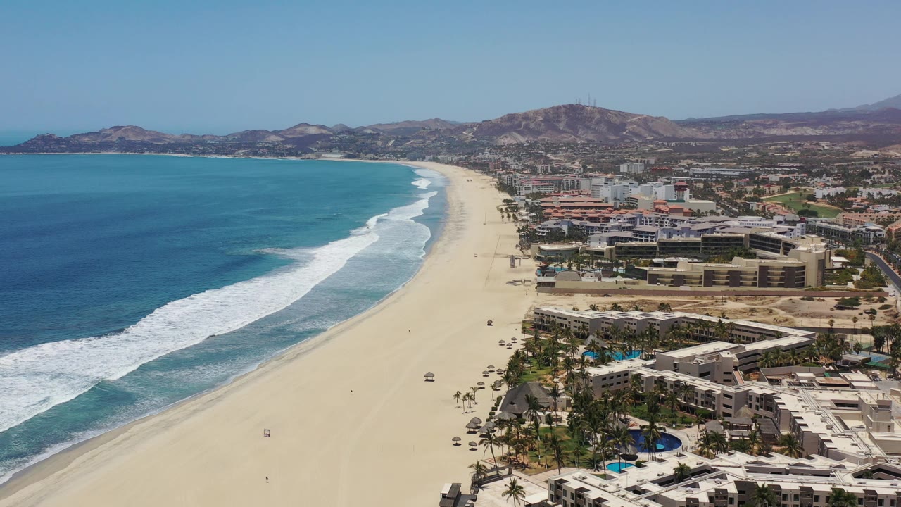 High aerial frame of the Los Cabos shoreline with rolling surf closer to the lower left, a long empty-looking sand strip, beachfront buildings, palms, pools, and no visible people.
