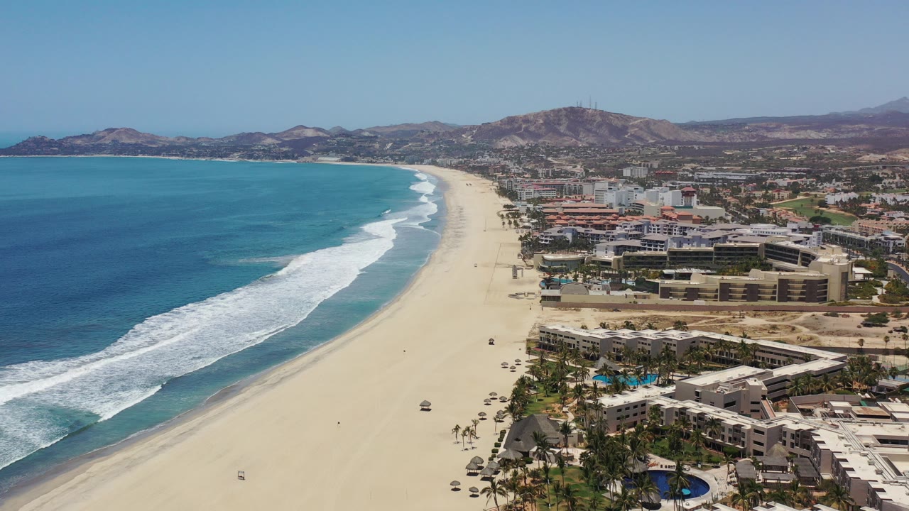 Opening aerial frame over Los Cabos, Mexico, with turquoise water on the left, a long pale beach, beachfront hotels and palms on the right, distant dry mountains, and no visible people.
