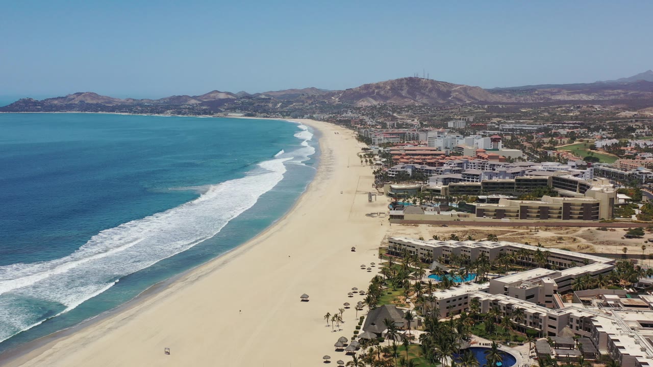 Oblique drone view of ocean waves beside a long beach and beachfront hotels.