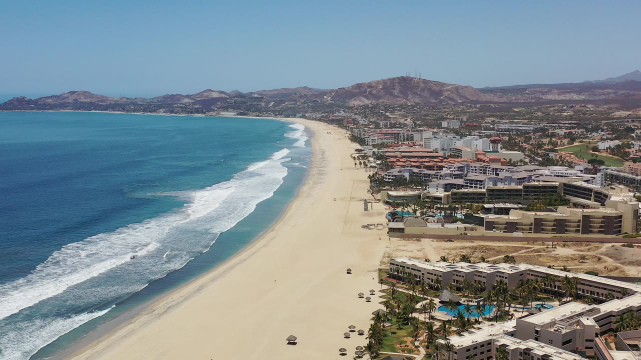 Aerial view of beach, blue ocean, surf, resort buildings, and hills.