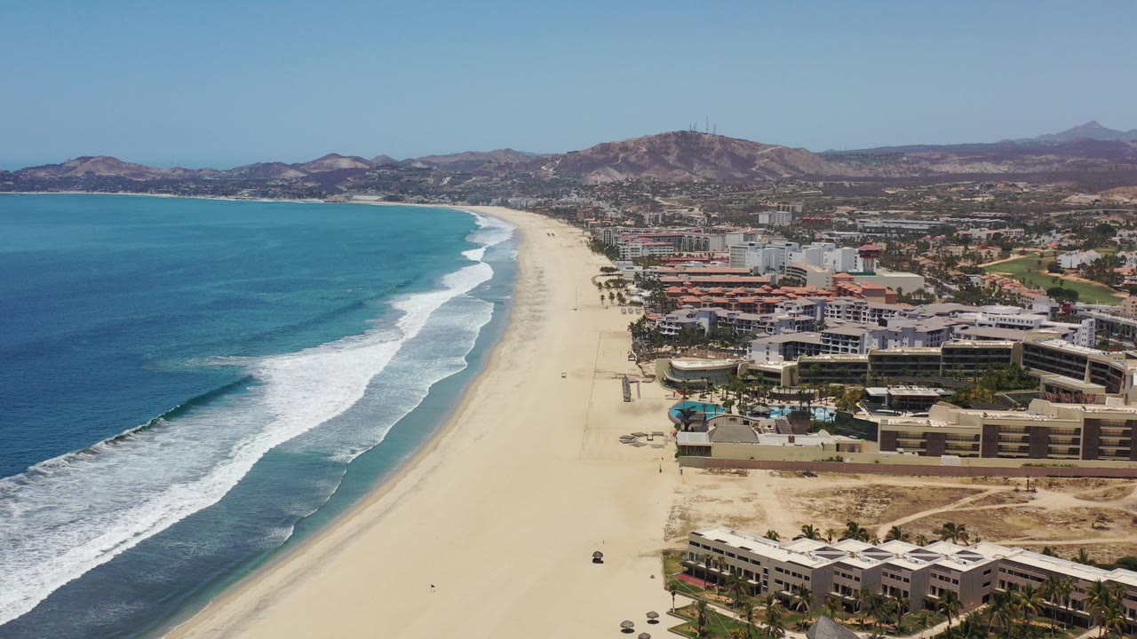 High aerial frame over the Los Cabos coastline with blue ocean at left, white surf on a wide sandy beach, resort buildings along the shore, inland hills beyond, and no visible people.