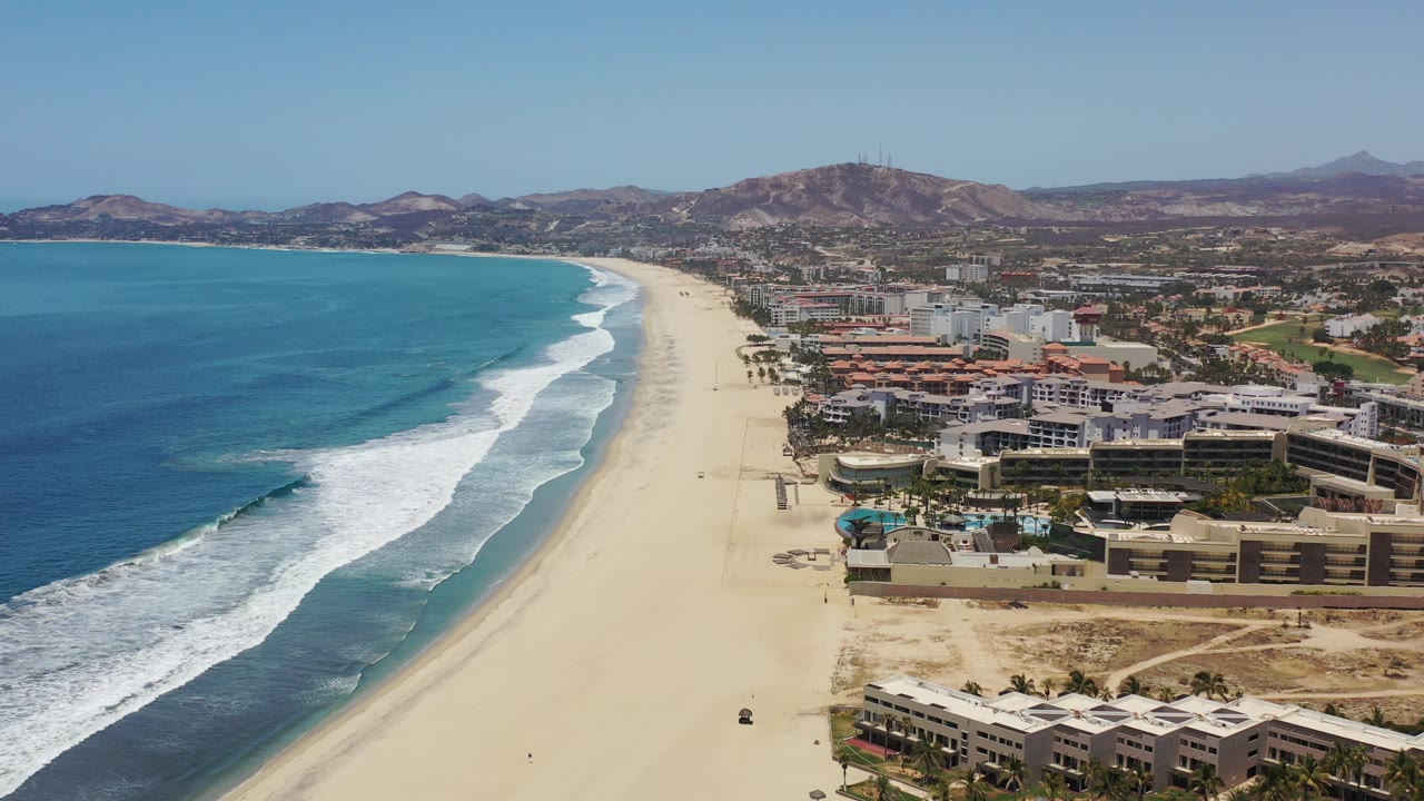 Los Cabos aerial frame showing turquoise water and white foam at left, a pale sand beach through the center, resort complexes and roads to the right, distant hills, and no visible people.