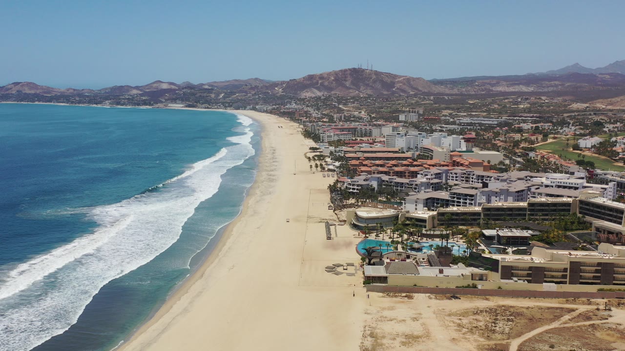 Aerial view of a wide sandy beach, ocean surf, shoreline buildings, and low hills.