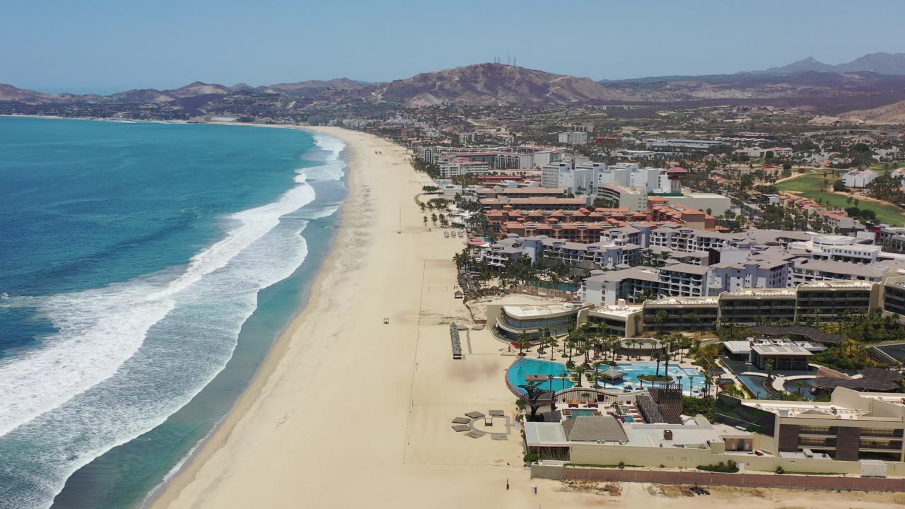 Opening aerial frame over the Los Cabos coastline, with turquoise water and surf on the left, a long sand beach in the center, beachfront buildings on the right, hills beyond, and no visible people.