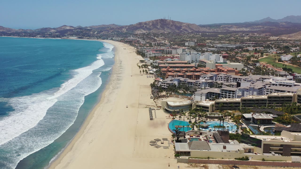 Drone view of Los Cabos beach curving past hotels and inland hills.